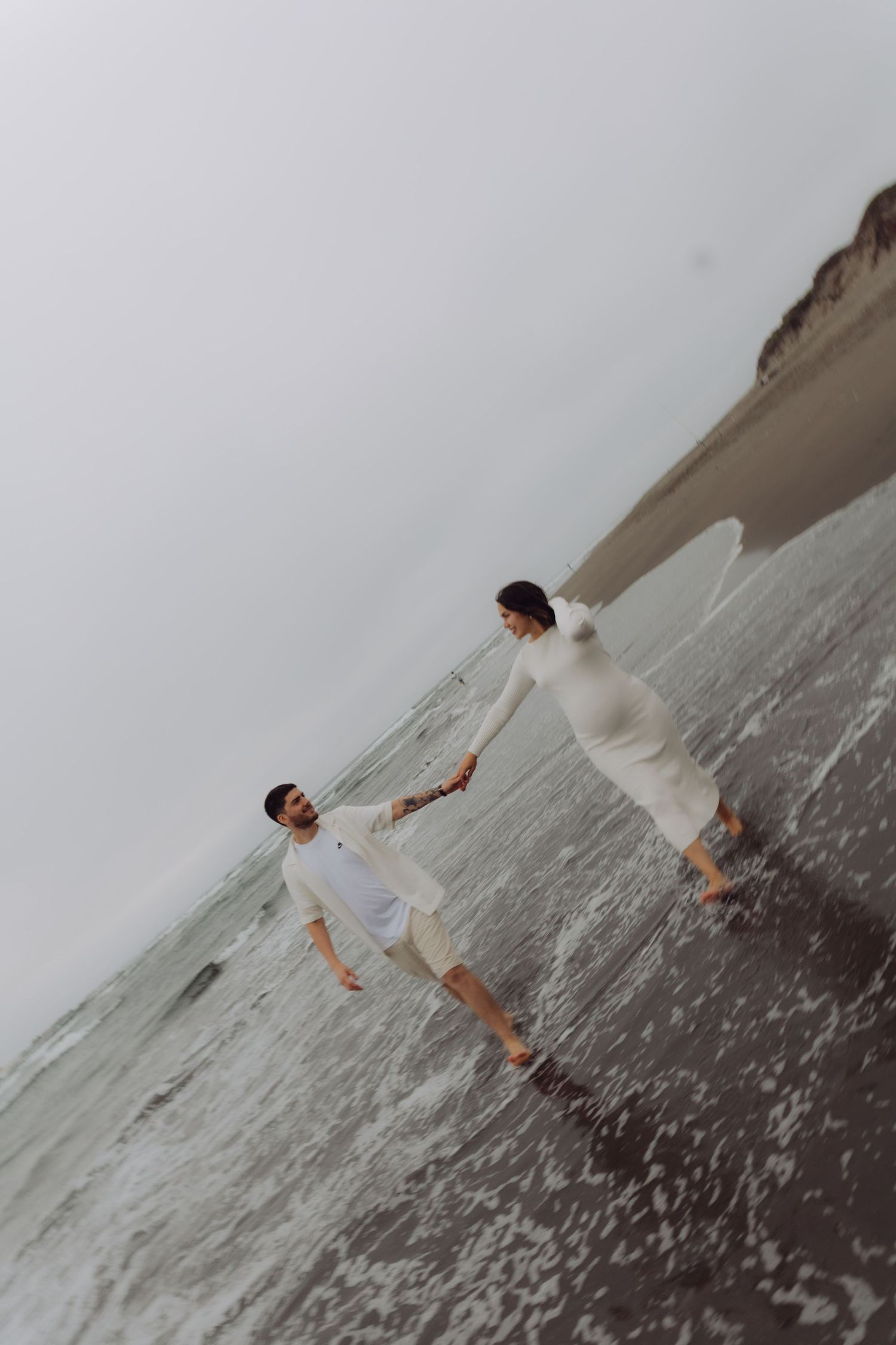 Expecting couple in matching white outfits holds hands while walking along a cloudy beach shoreline.