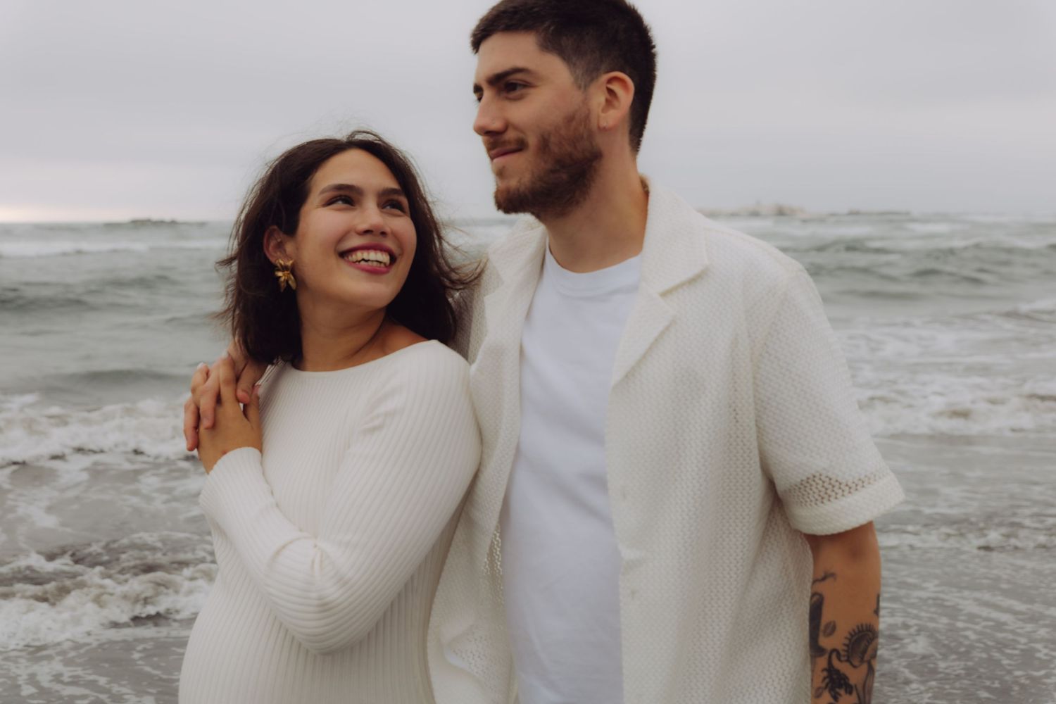 Happy couple in white outfits embracing on a beach with ocean waves behind them.