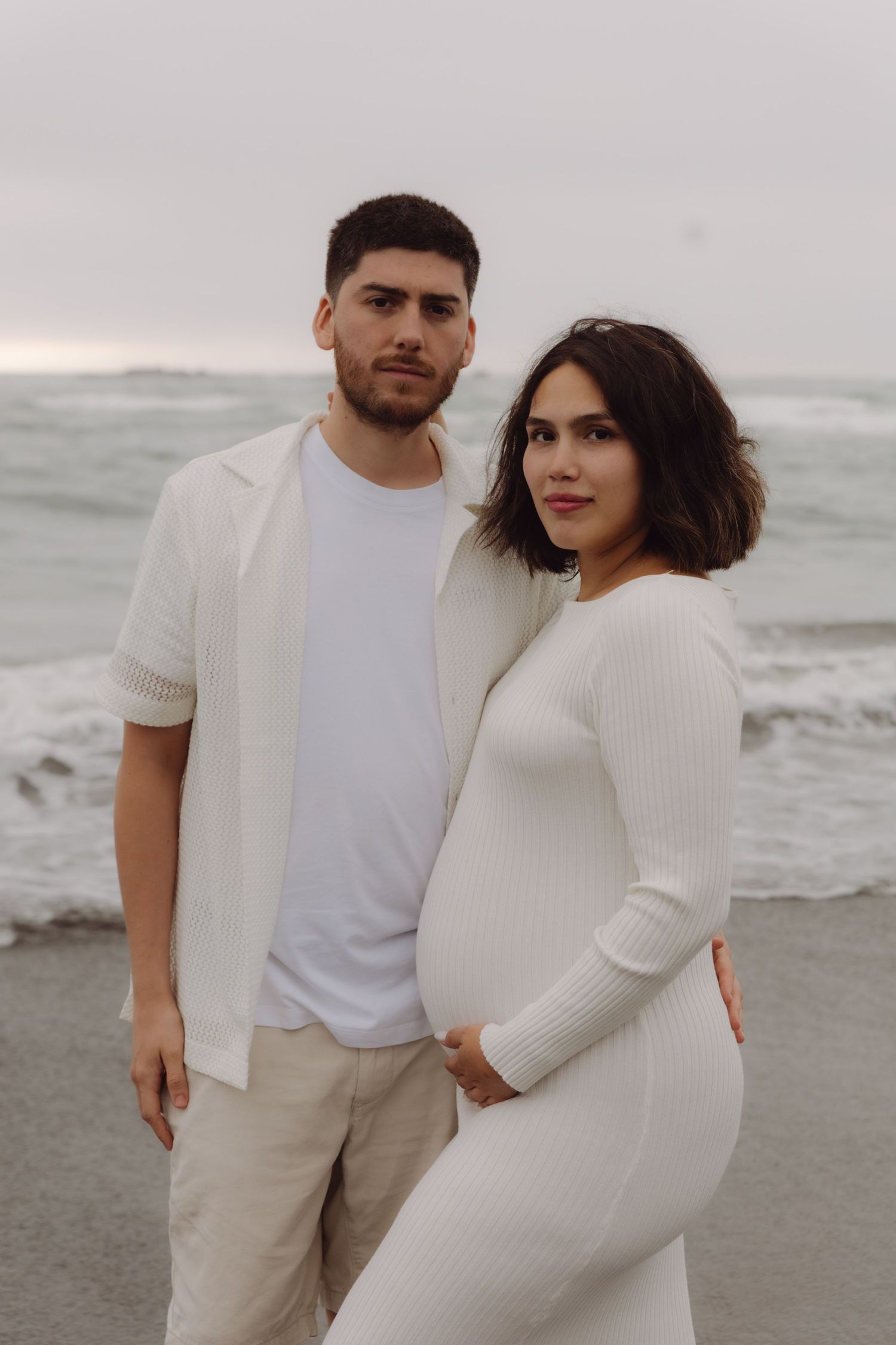 Pregnant couple in white outfits posing together on a cloudy beach.
