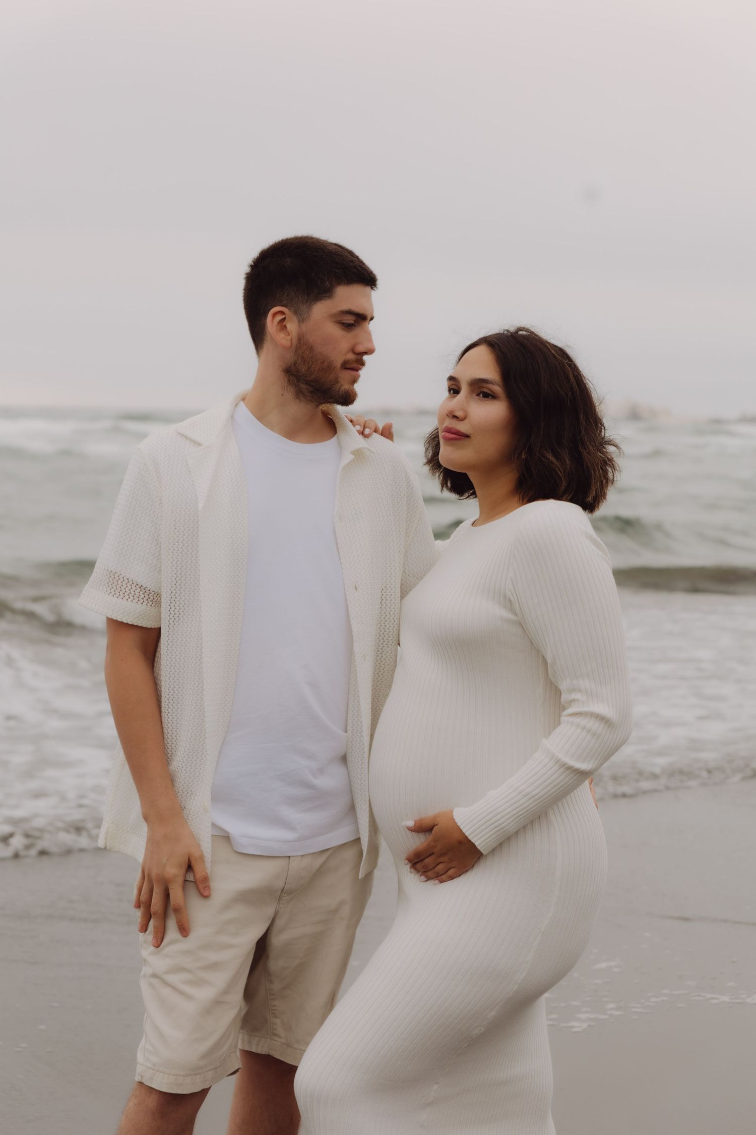 Expecting couple standing together on a beach, woman in white maternity dress.
