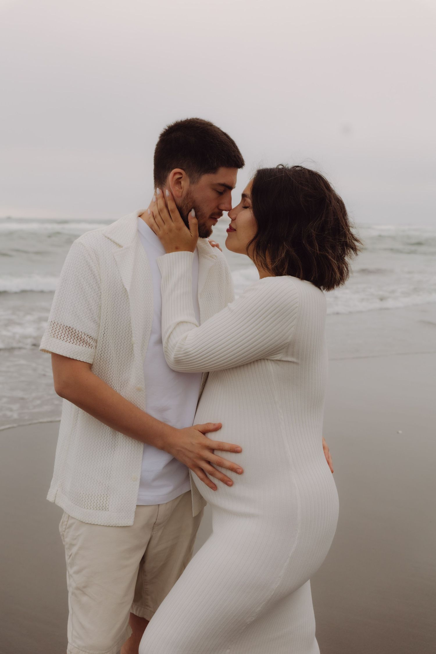 Pregnant couple sharing a kiss on the beach with ocean waves in background.