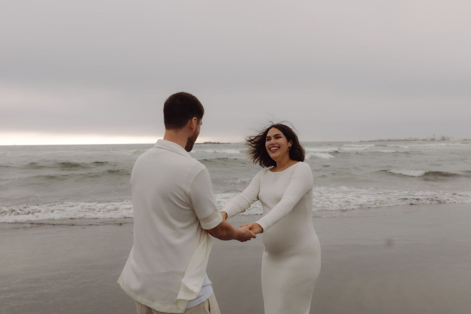 Smiling pregnant woman holding hands with partner on a windy beach.
