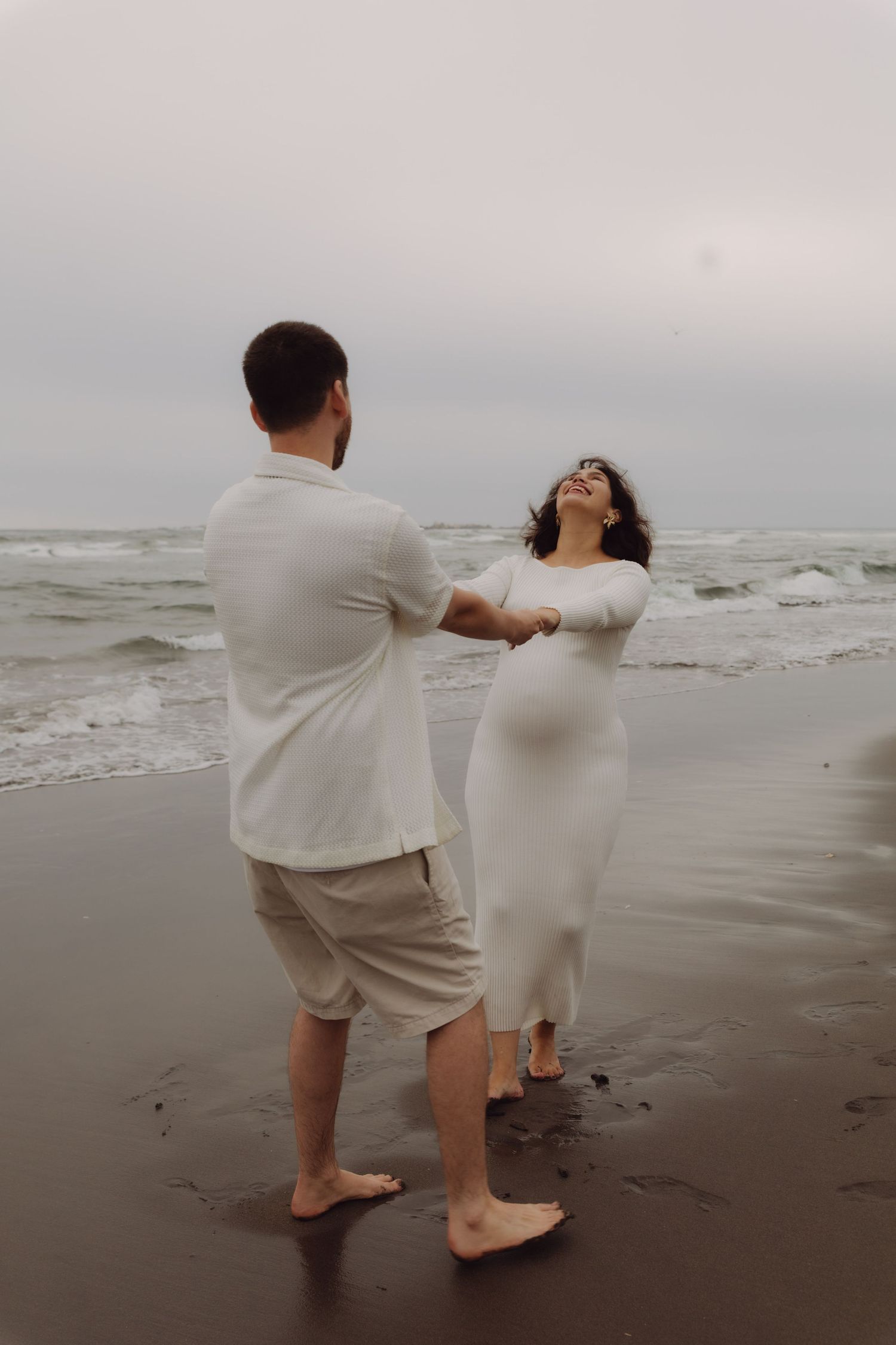 Barefoot expecting couple dancing and laughing together on a cloudy beach.