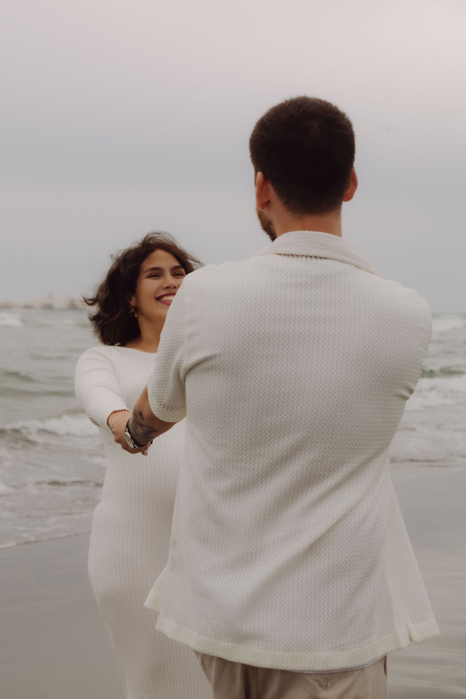 Laughing pregnant woman holding hands with partner on a cloudy beach.