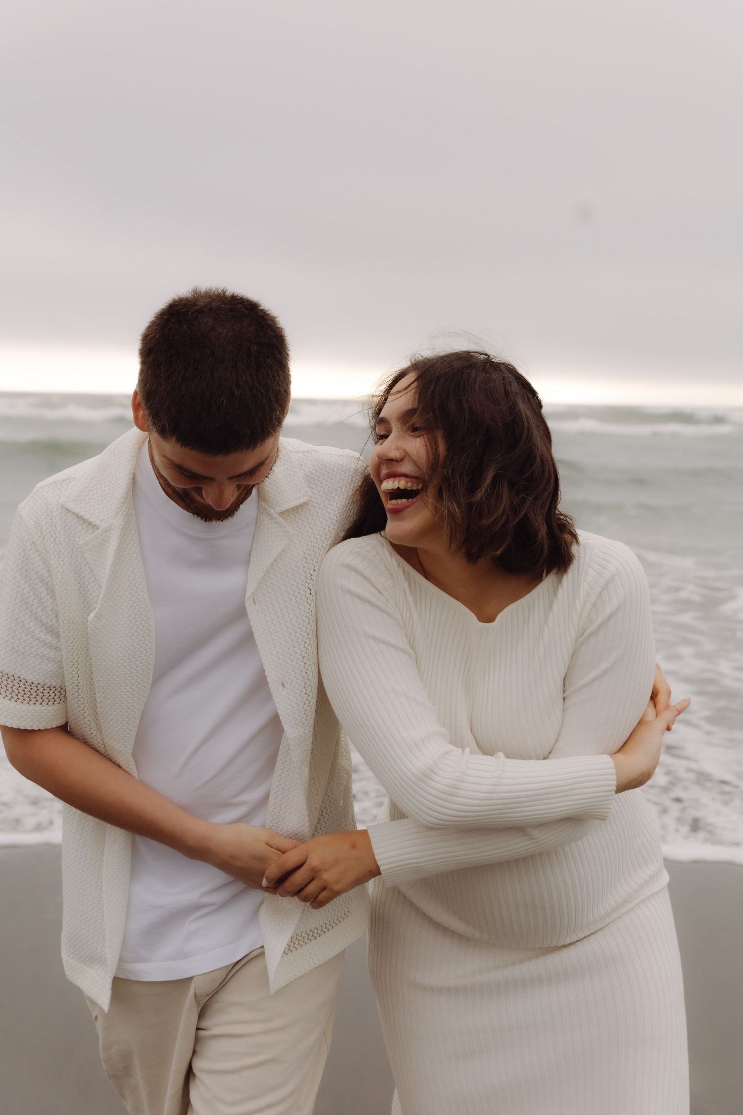 Joyful pregnant couple laughing and holding hands on a breezy beach.