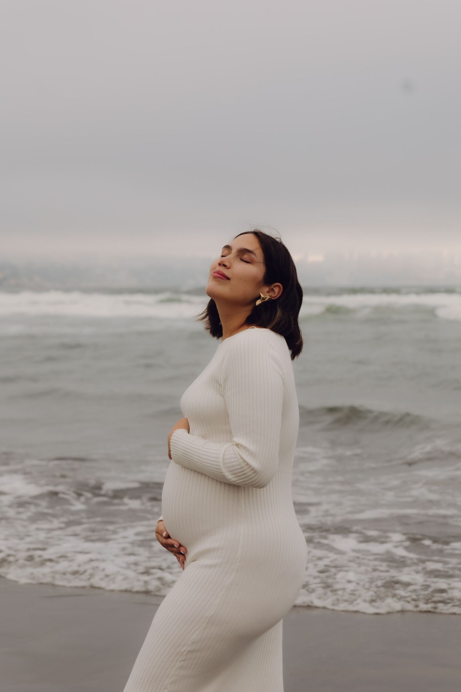 Pregnant woman in white ribbed dress stands on beach with eyes closed, holding baby bump near ocean waves.