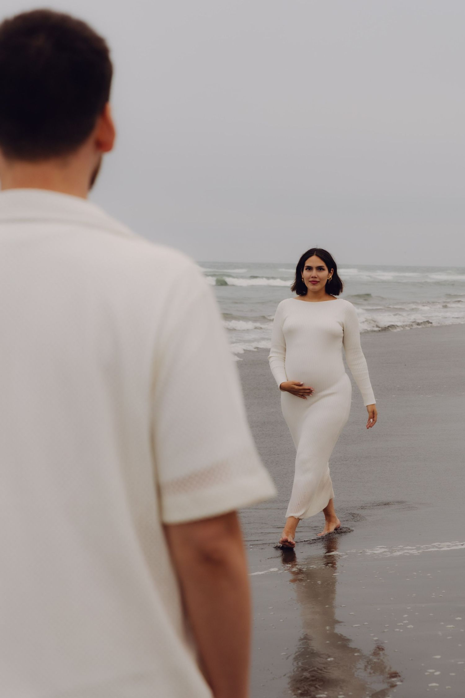 Pregnant woman in white dress walks barefoot on beach toward man in white shirt near ocean.