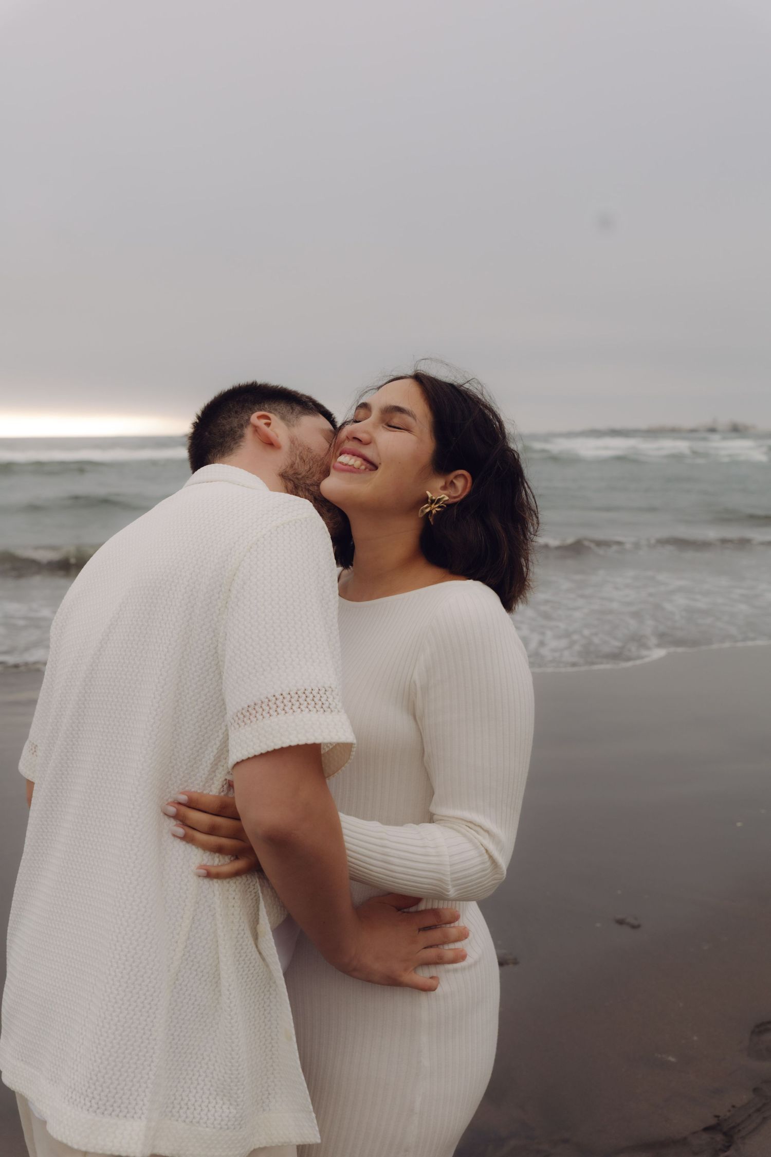 Couple embracing on beach, man kissing pregnant woman's cheek as she smiles, both wearing white.