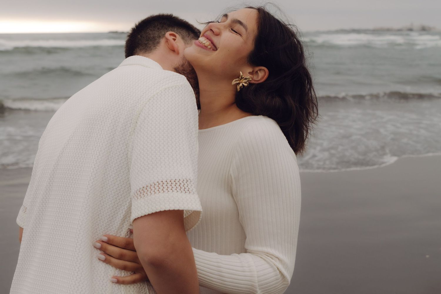 Man kisses smiling pregnant woman on beach, both in white outfits with ocean waves behind them.