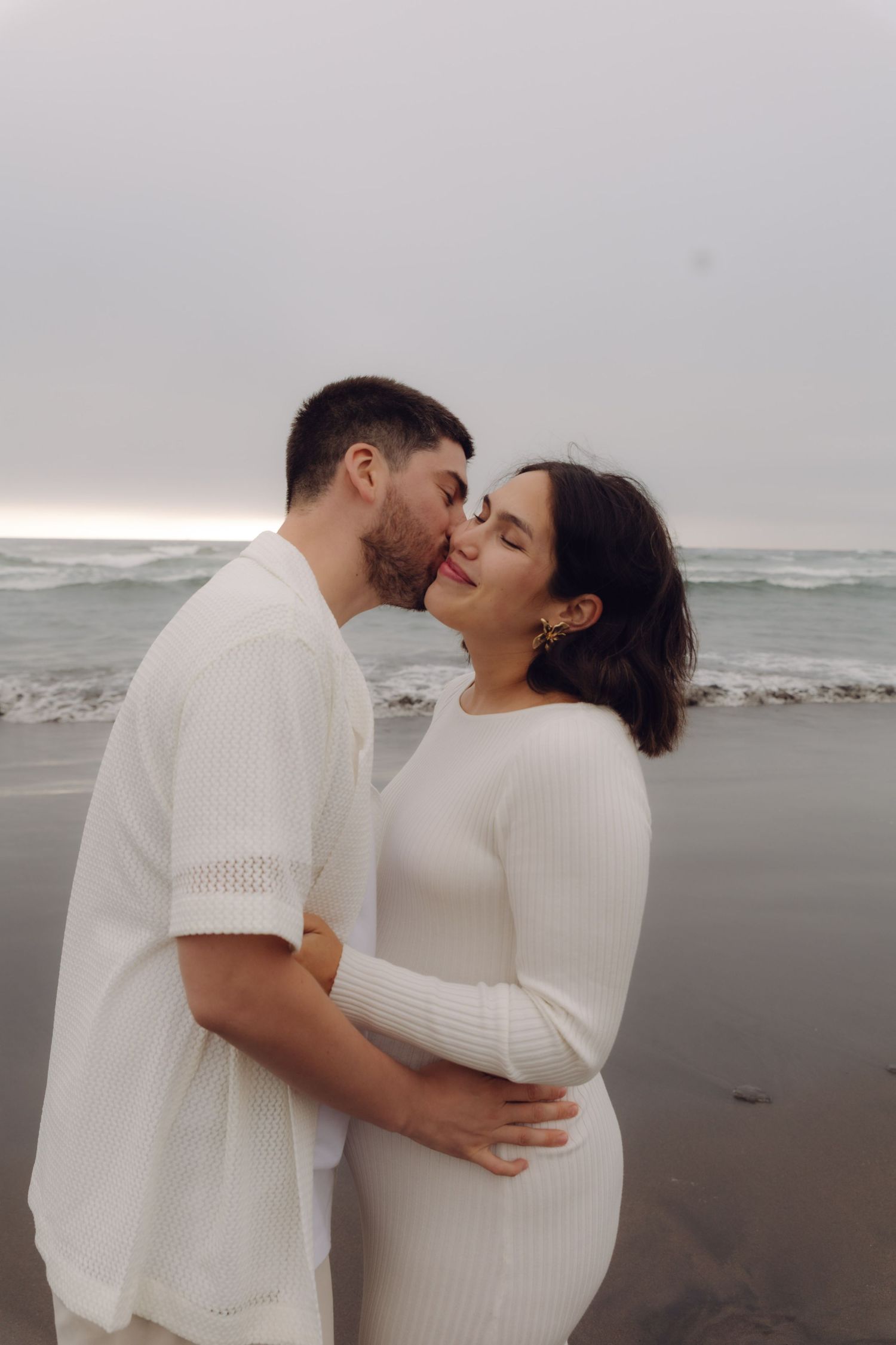 Couple sharing a kiss on a cloudy beach, man embracing pregnant woman in white ribbed dress.
