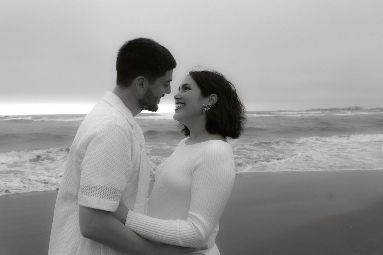 Black and white photo of couple embracing and smiling at each other on a beach.