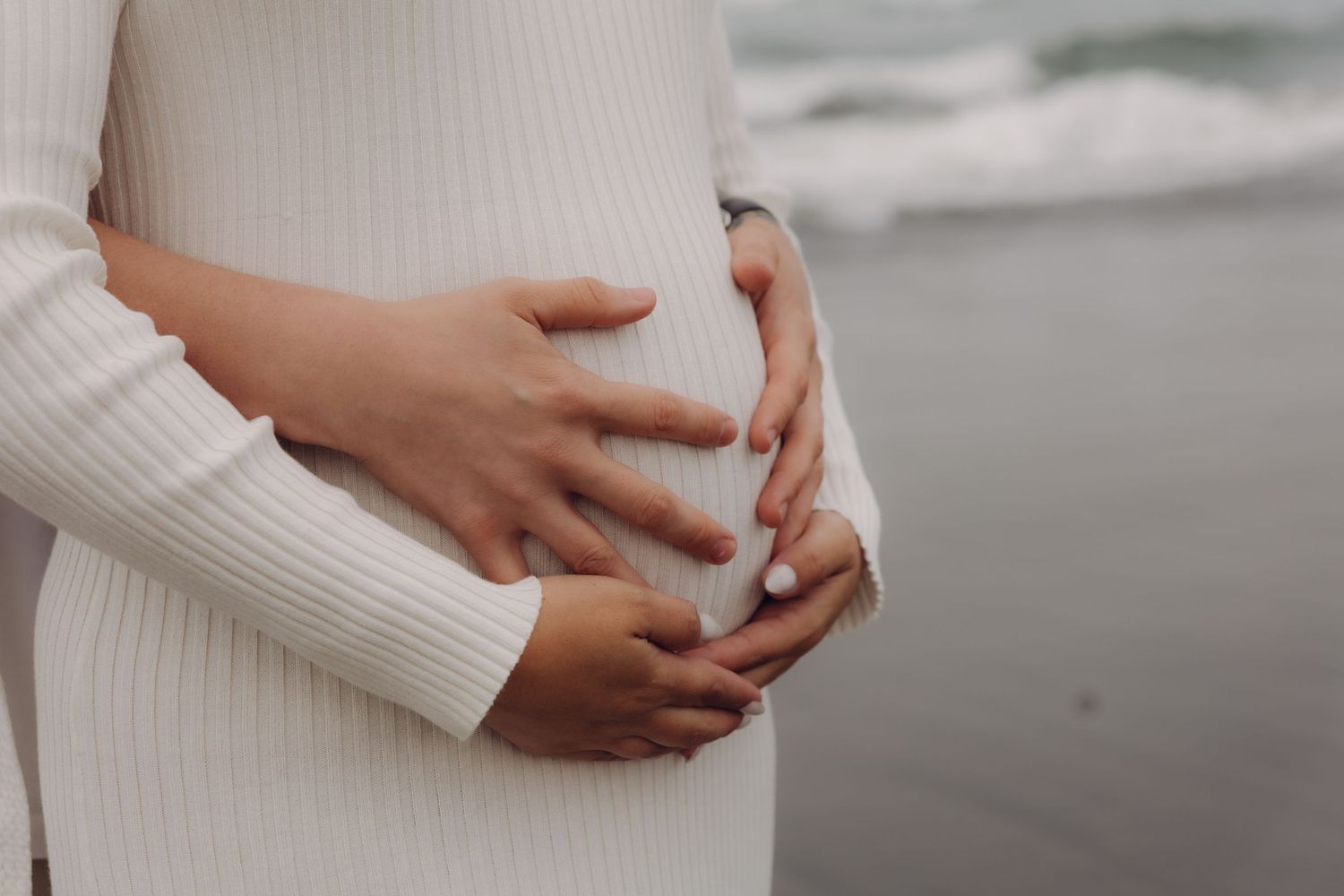 Close-up of couple's hands cradling pregnant belly in white ribbed dress near ocean shore.