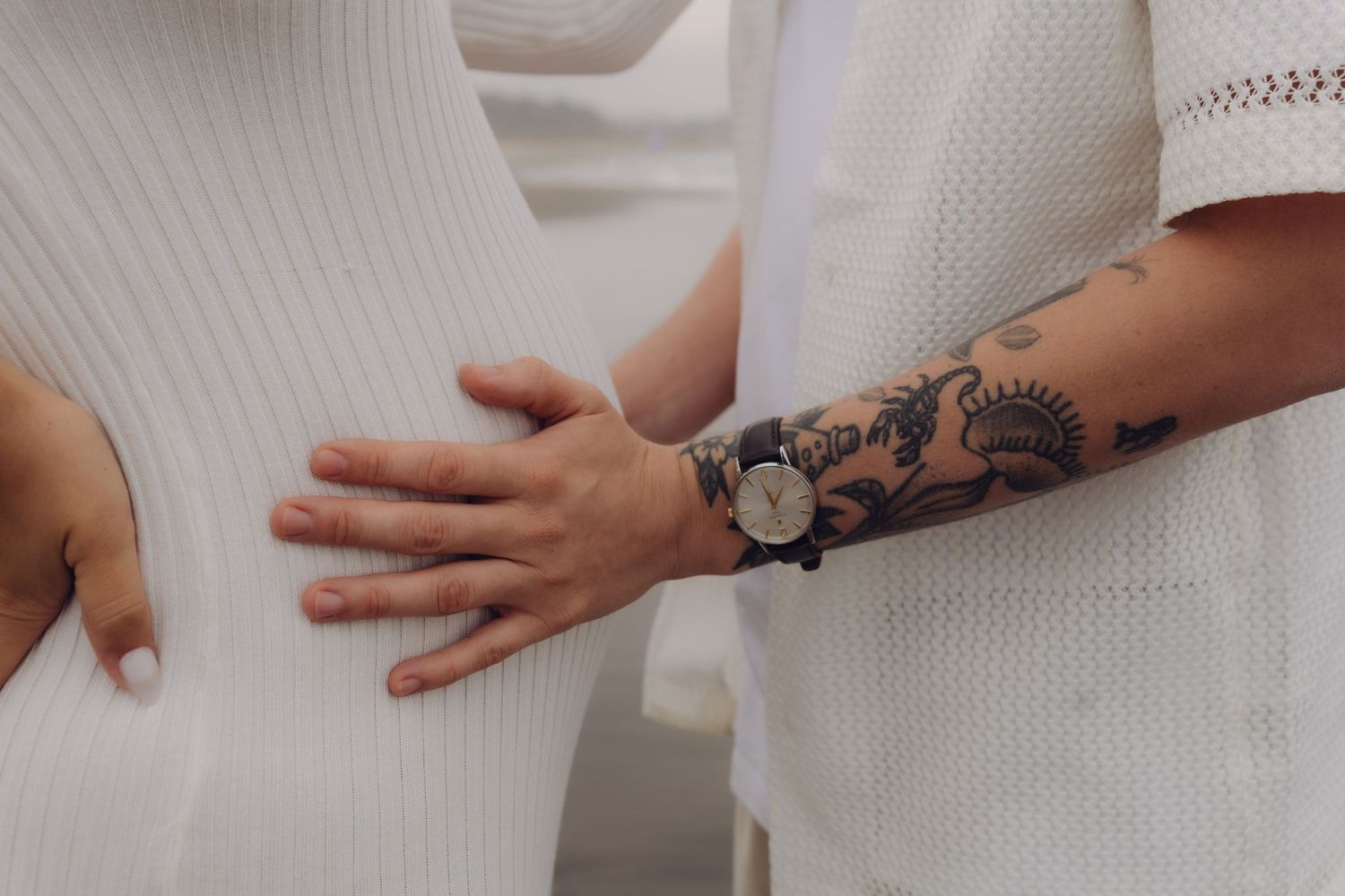Close-up of tattooed man's hand resting on pregnant woman's white ribbed dress belly.