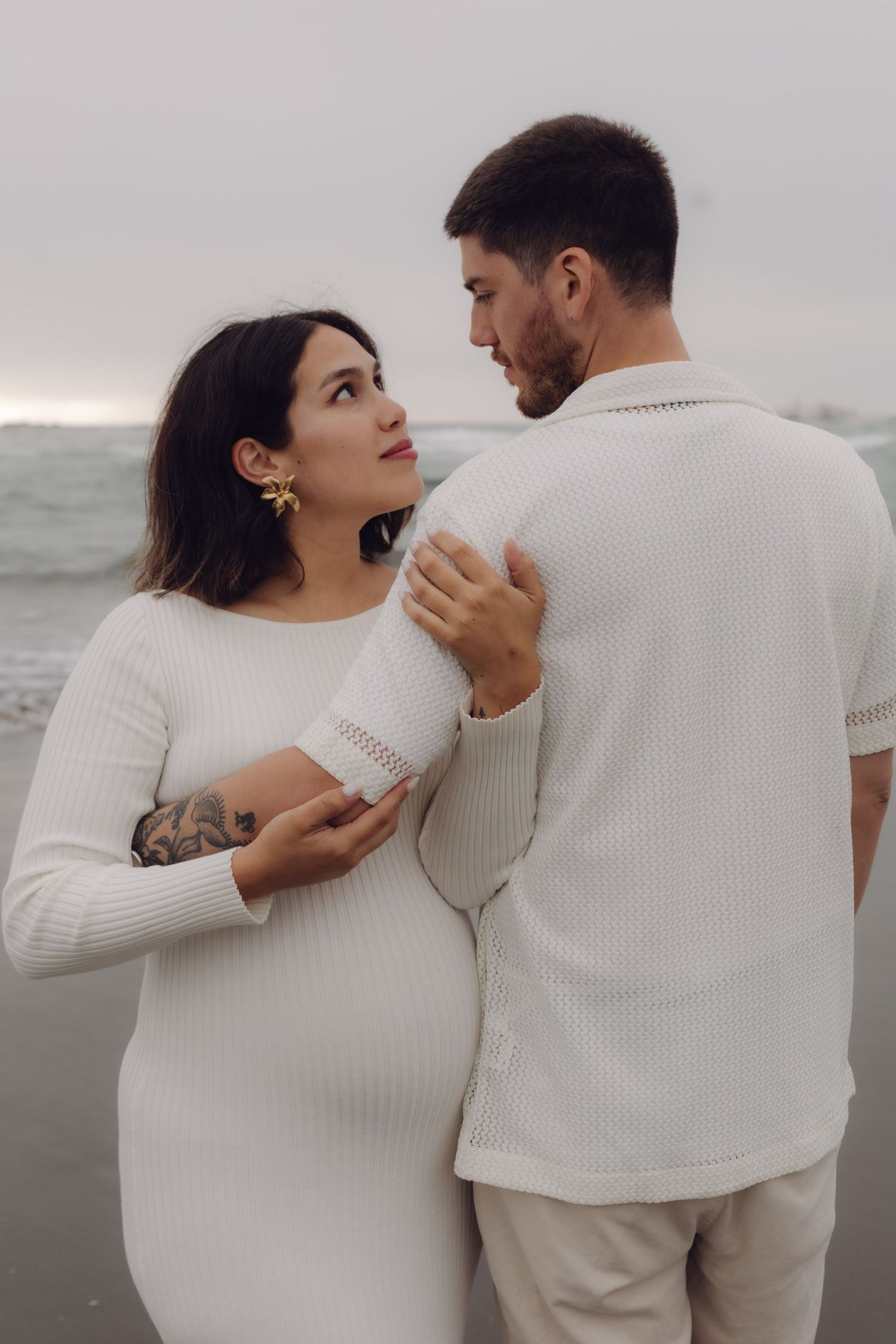 Pregnant woman with tattoo and gold earrings gazes at partner on overcast beach, both in white.