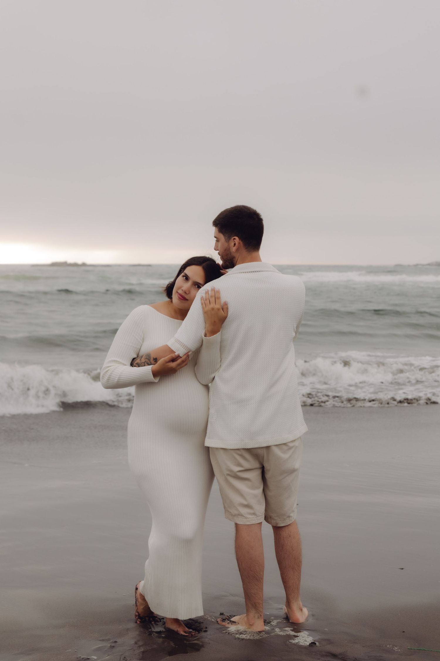 Pregnant woman in white dress leans on partner's shoulder as they stand together on ocean beach.