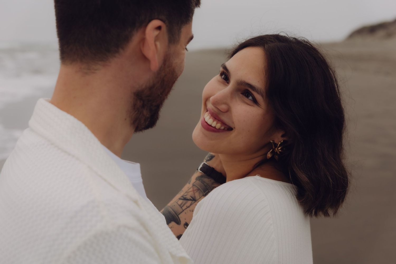 Smiling woman with dark hair and tattoos embraces man on a misty beach during a maternity photoshoot.