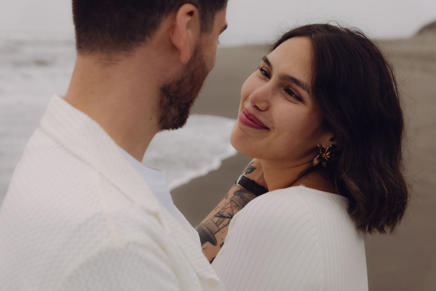 Couple sharing an intimate moment on a cloudy beach, woman with tattoos smiling warmly at her partner.