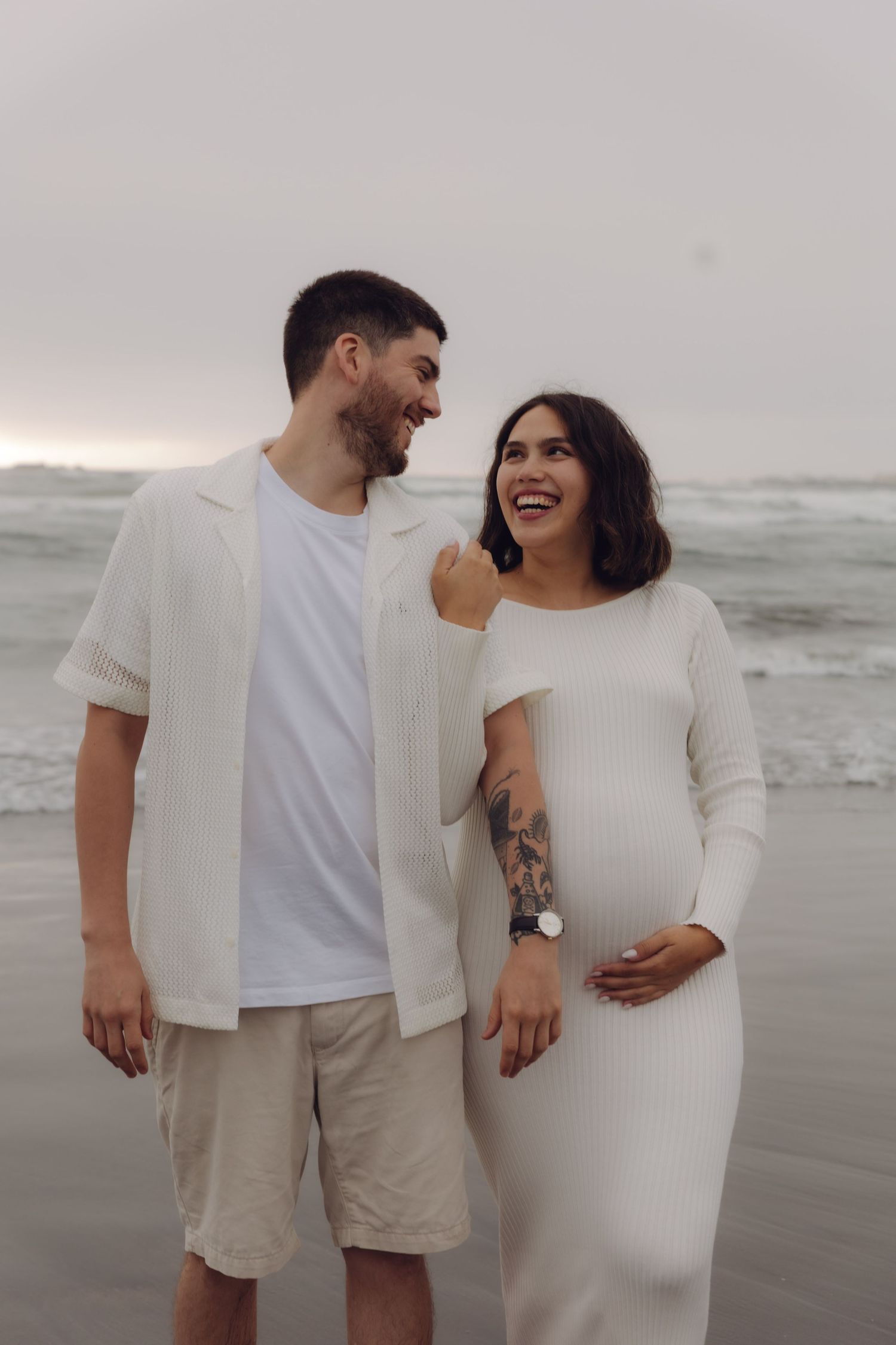 Happy expecting couple in white outfits laughing together on a misty beach during maternity shoot.