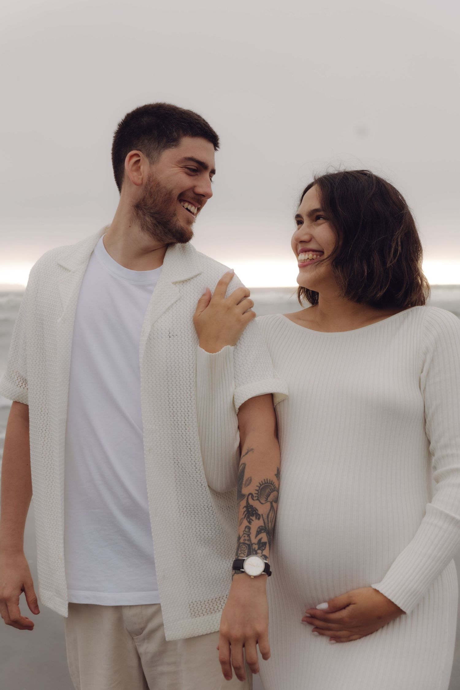 Smiling pregnant woman in white ribbed dress holds baby bump while standing with partner on beach.