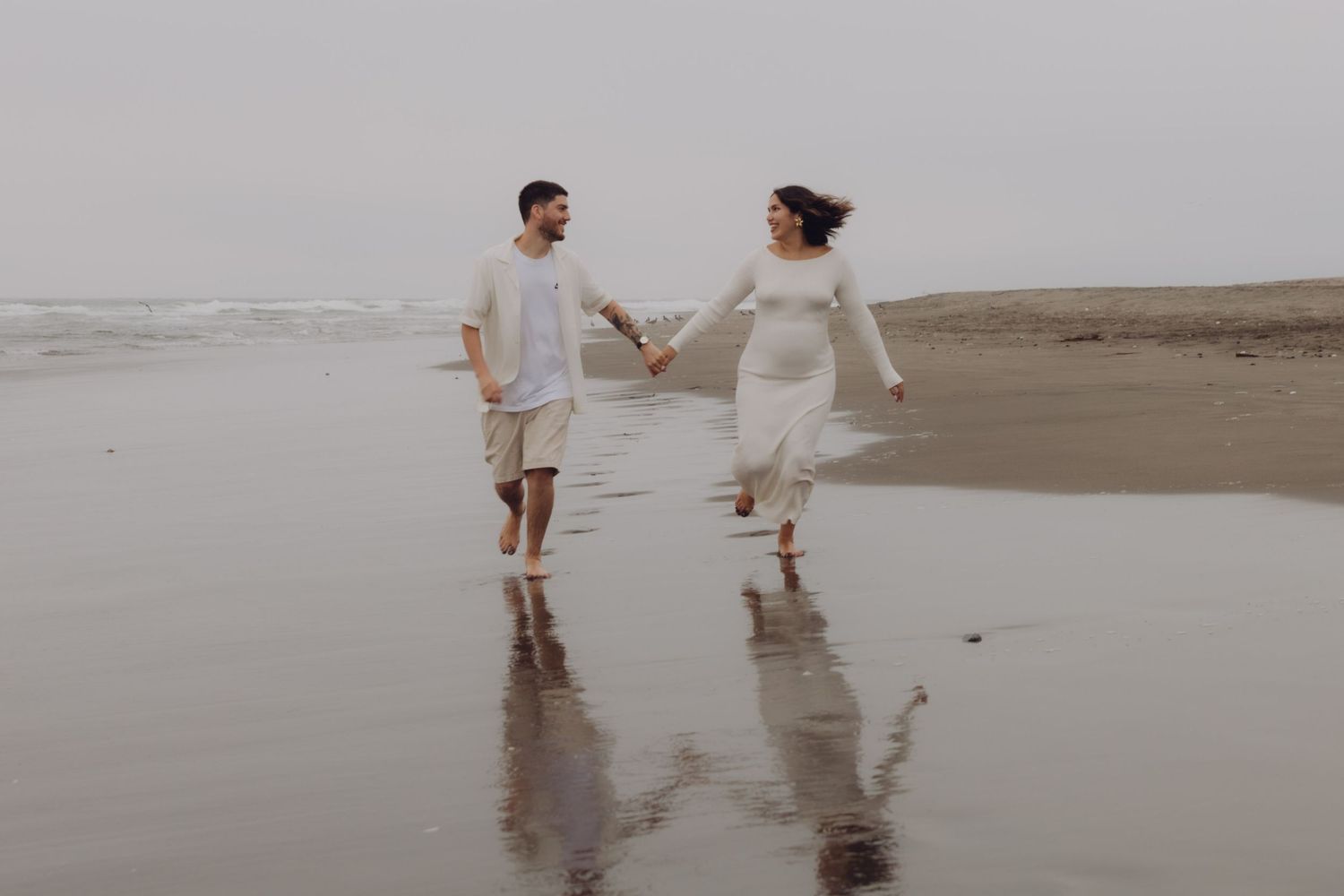Expecting couple holding hands and walking along a reflective wet beach shore on an overcast day.