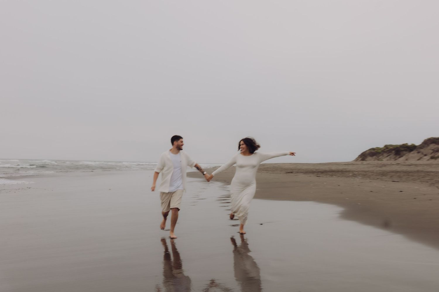 Joyful pregnant couple in white outfits dancing and holding hands on a foggy beach shoreline.