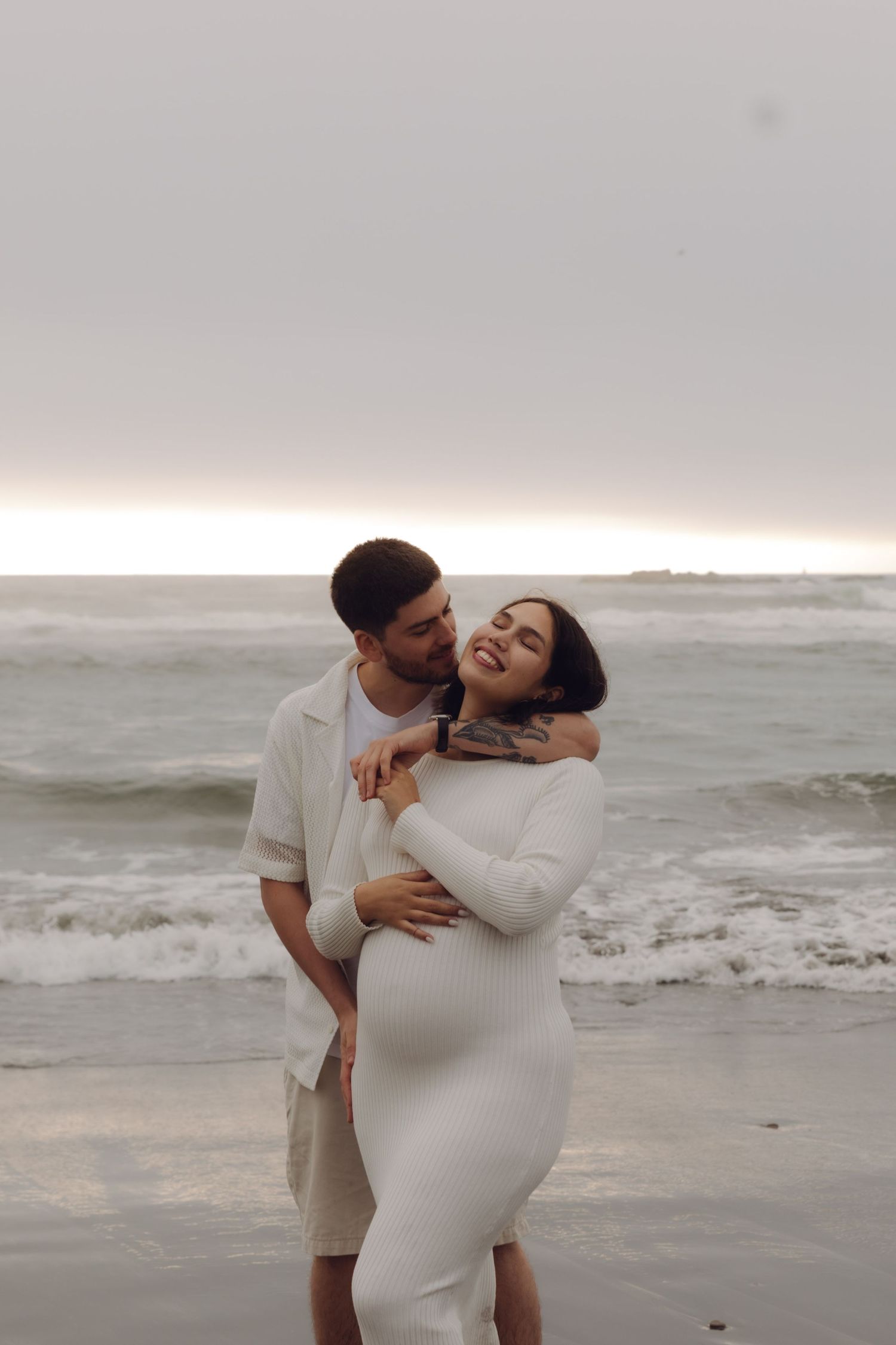 Man embraces smiling pregnant woman in white fitted dress from behind on a scenic ocean beach.