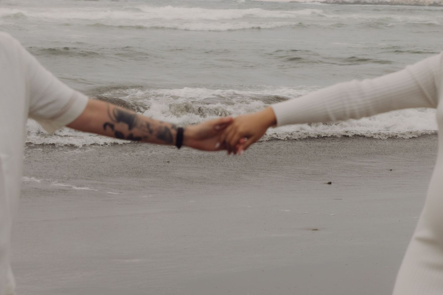 Close-up of tattooed couple holding hands near ocean waves during a romantic beach maternity session.
