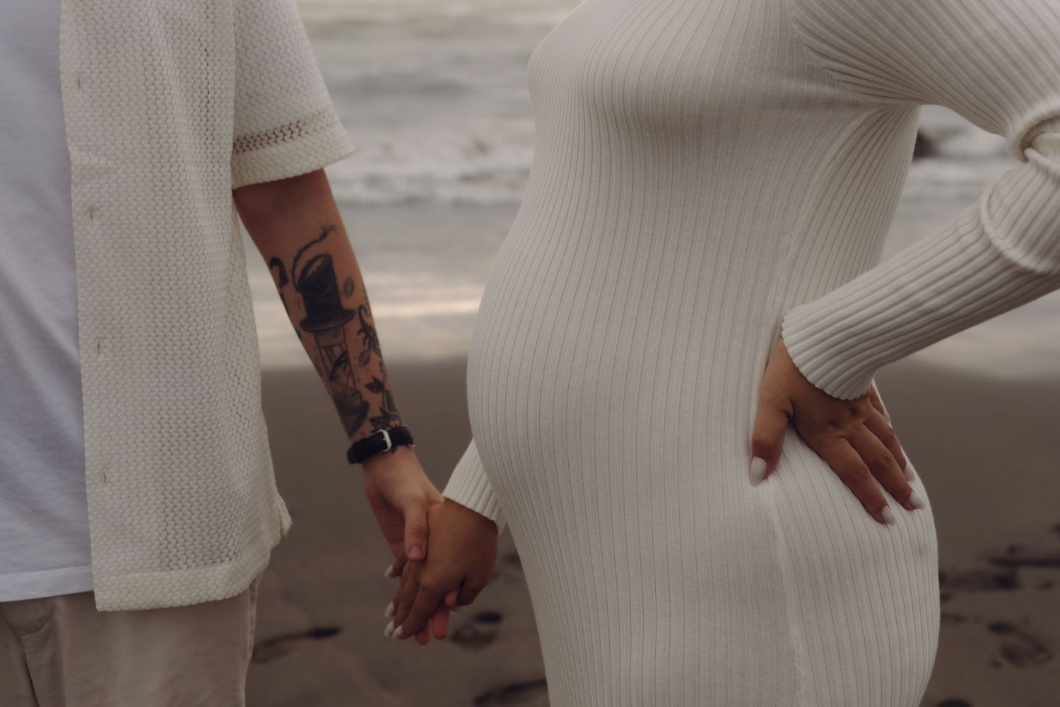 Close-up of pregnant woman in white ribbed dress holding hands with tattooed partner on beach.