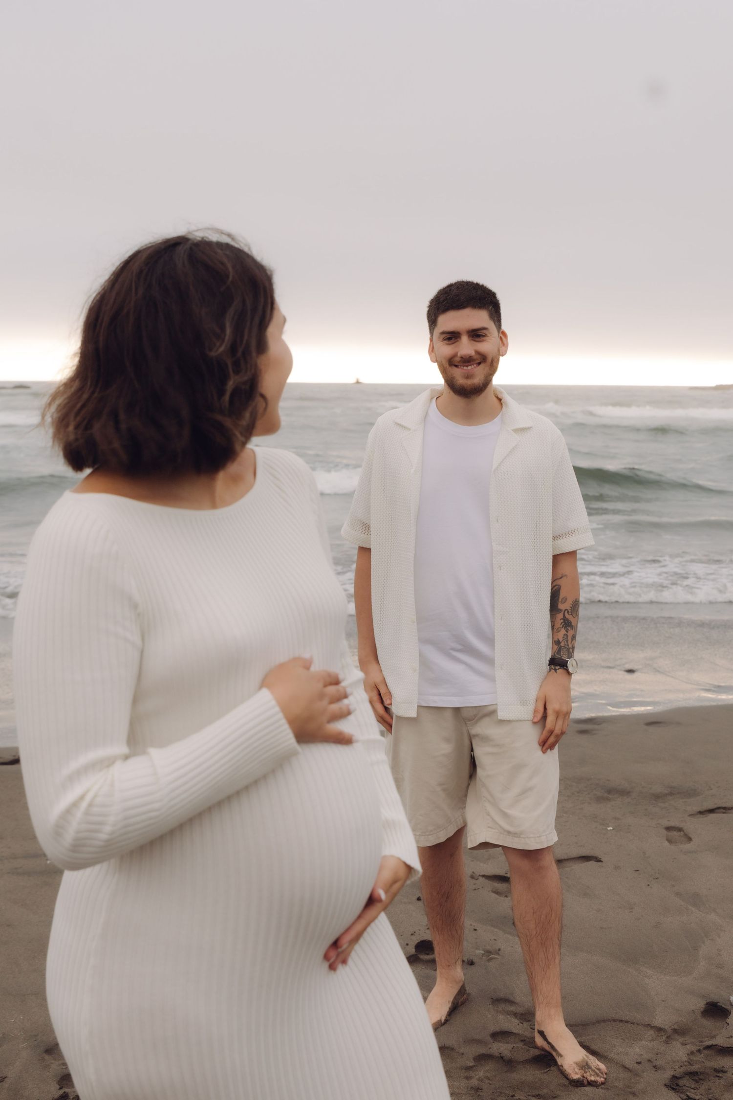 Pregnant woman in white ribbed dress holds baby bump as smiling partner stands on misty beach.