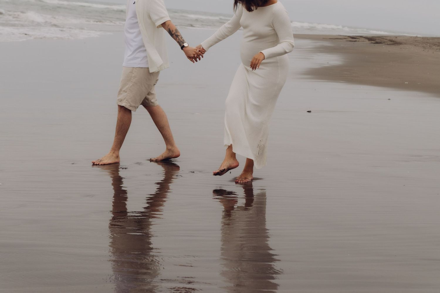 Pregnant couple holding hands walking barefoot on a dark sand beach near the ocean waves.