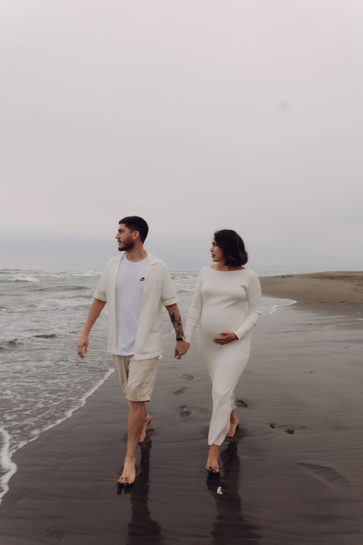 Expecting couple walking hand in hand along a black sand beach with ocean waves behind them.