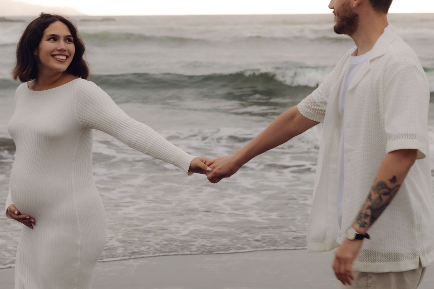 Smiling pregnant woman holding hands with tattooed partner on a beach with waves in background.