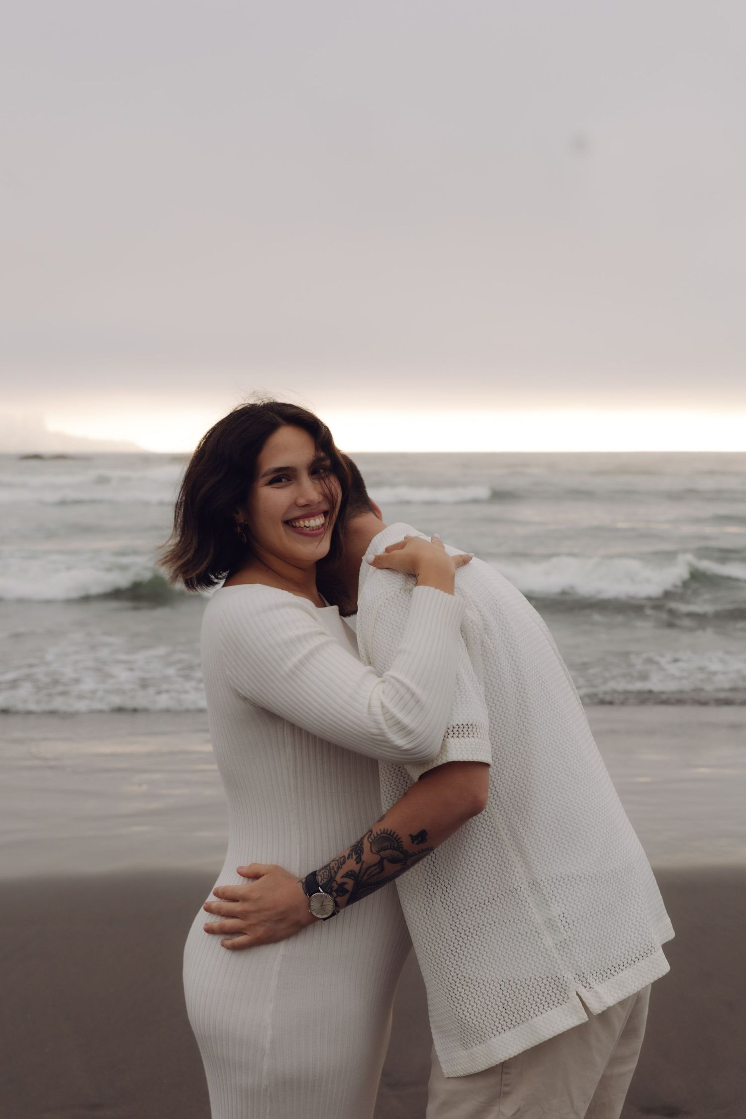 Pregnant woman in white dress smiling while embracing tattooed partner on a cloudy beach.