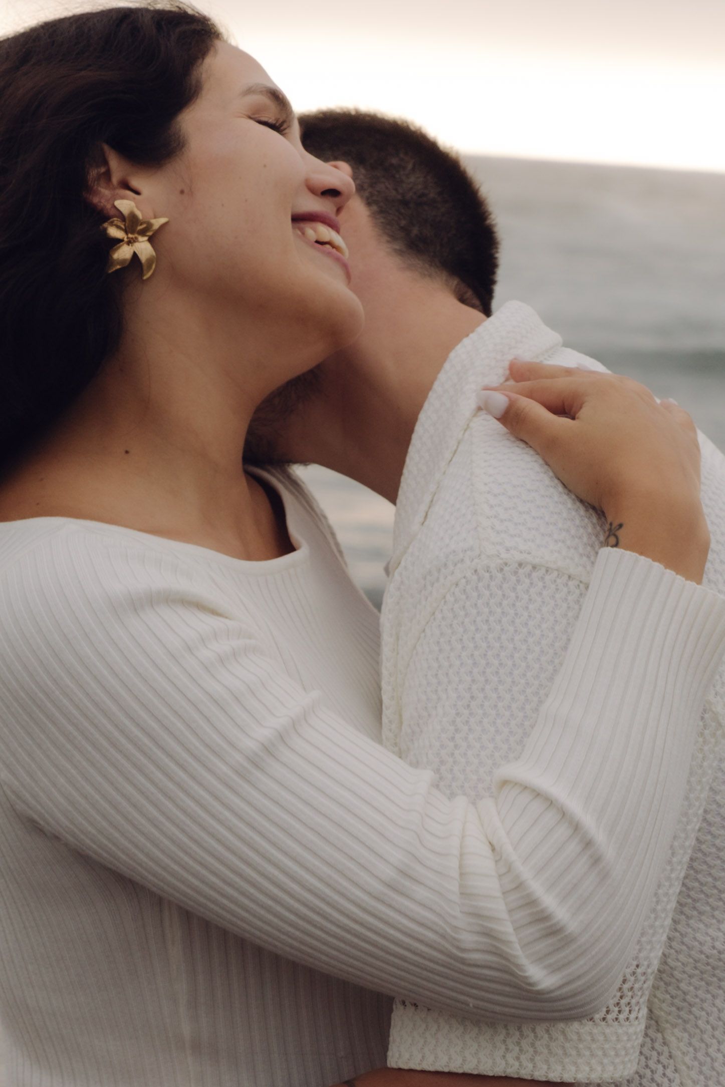 Close-up of smiling pregnant woman with gold earrings embracing partner on a misty beach.