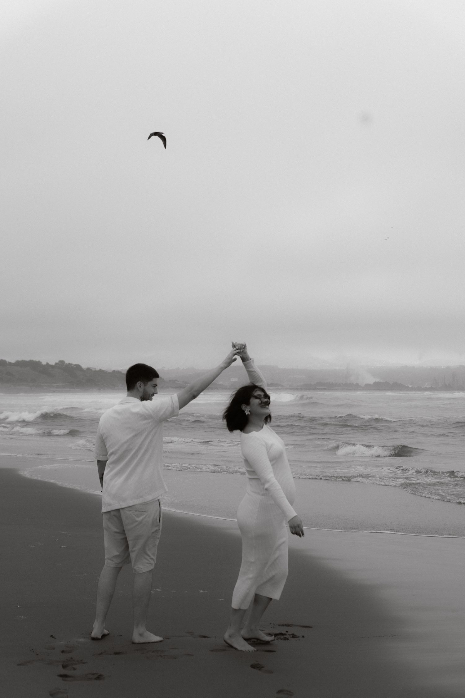 Black and white photo of couple dancing on beach with man spinning woman near ocean waves.
