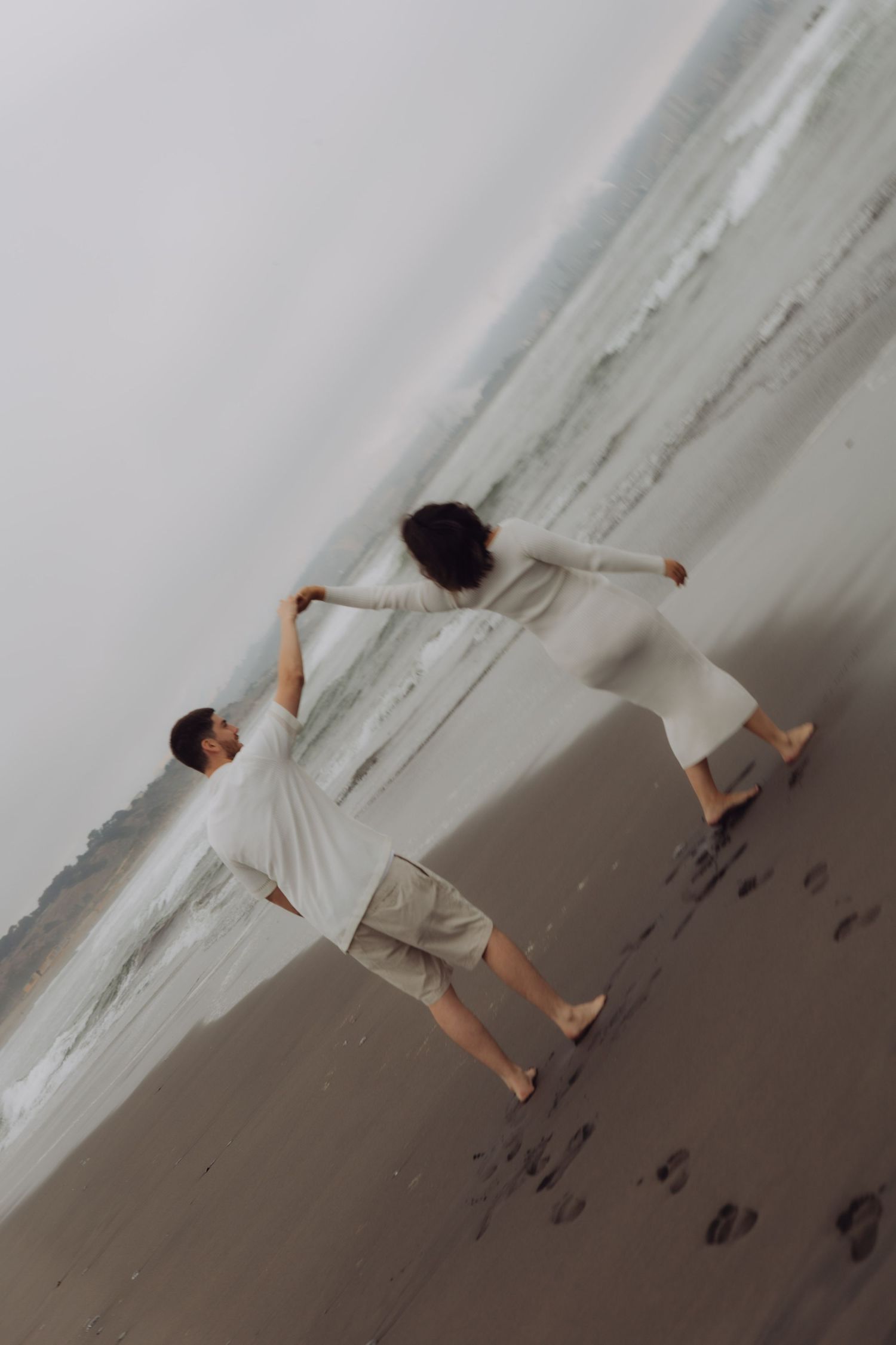 Couple holding hands and leaning away from each other playfully on a sandy beach shoreline.