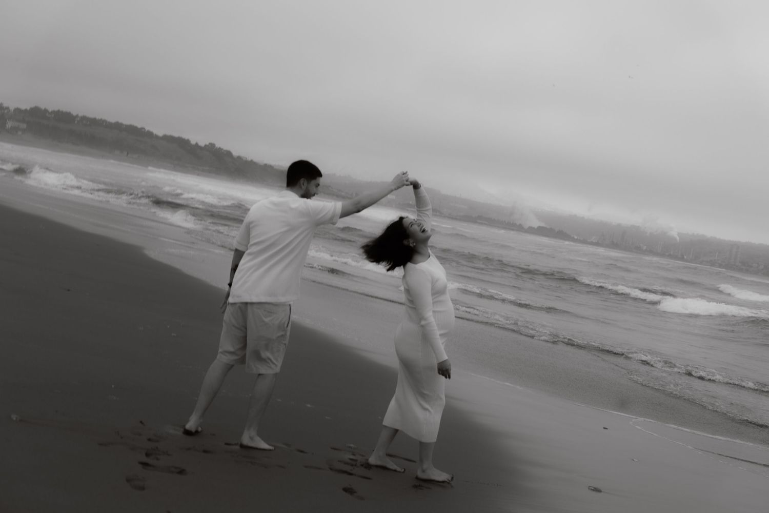 Black and white image of couple dancing on foggy beach with man twirling woman by the ocean.