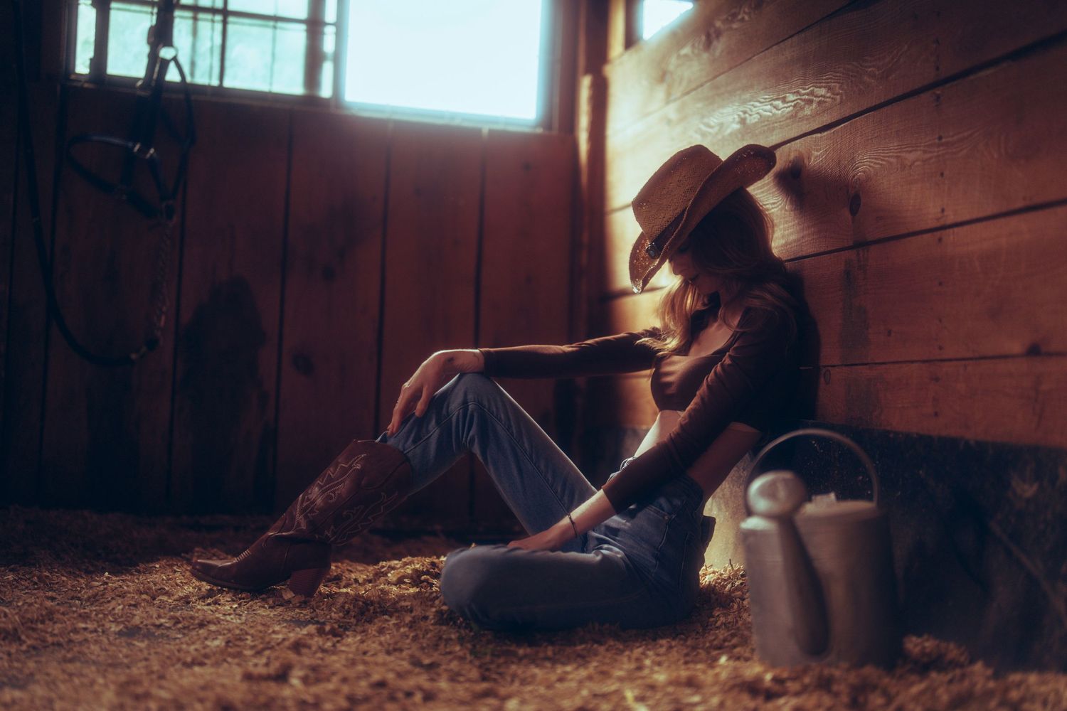 Person in cowboy hat rests in hazy sunlit barn with bucket nearby on hay-covered floor.