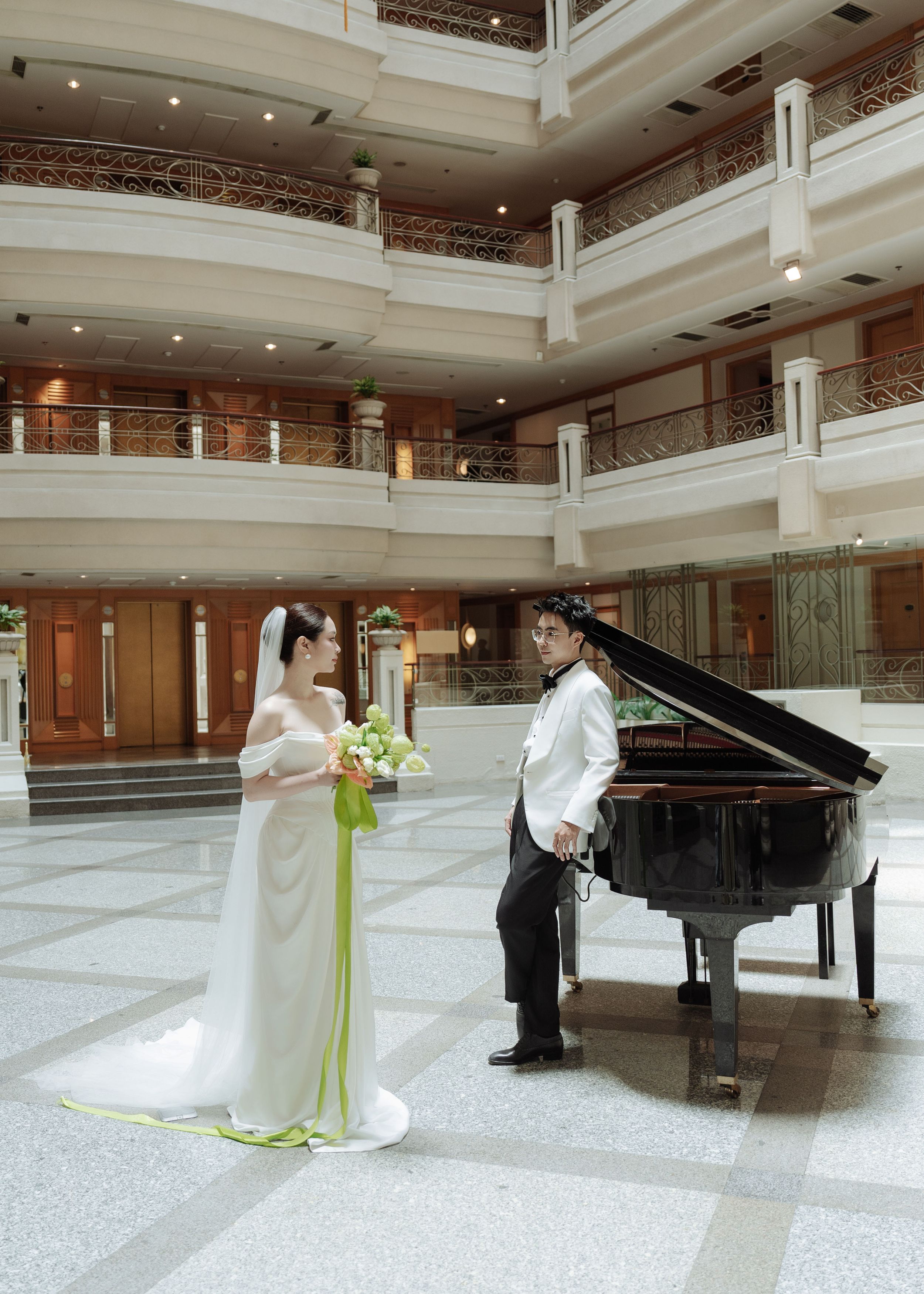 Bride and groom posing with a grand piano in an elegant multi-story hotel atrium with ornate balconies.