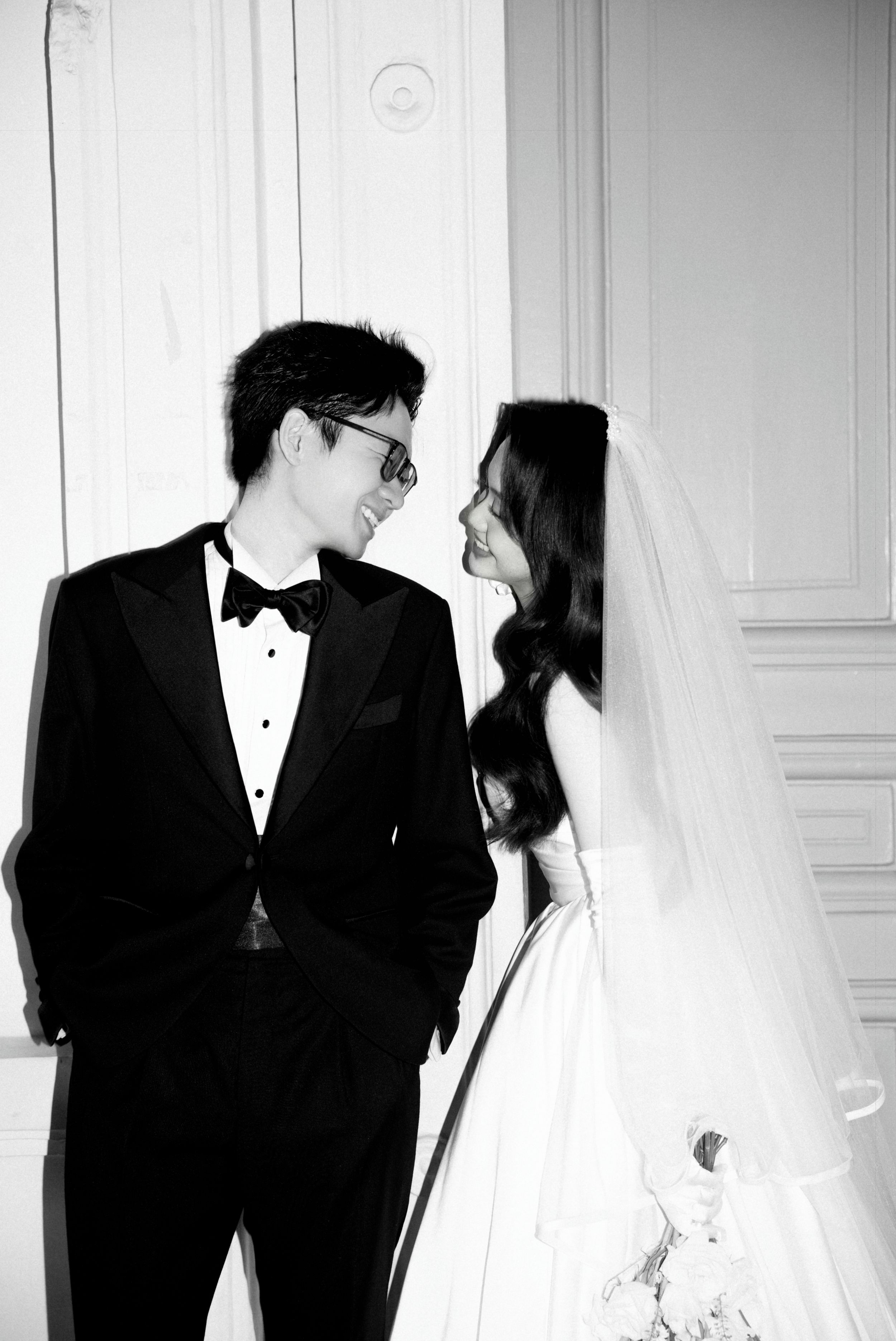 Black and white photo of smiling newlyweds in formal wedding attire gazing at each other indoors.