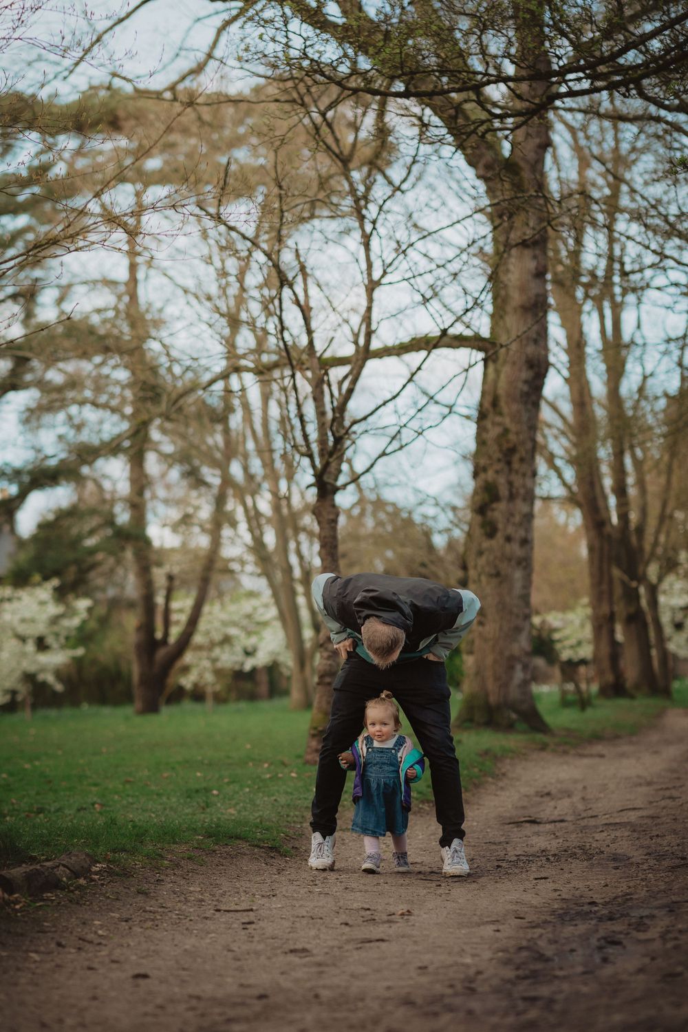 Adult and toddler walking together on a dirt path in a wooded park setting during spring.