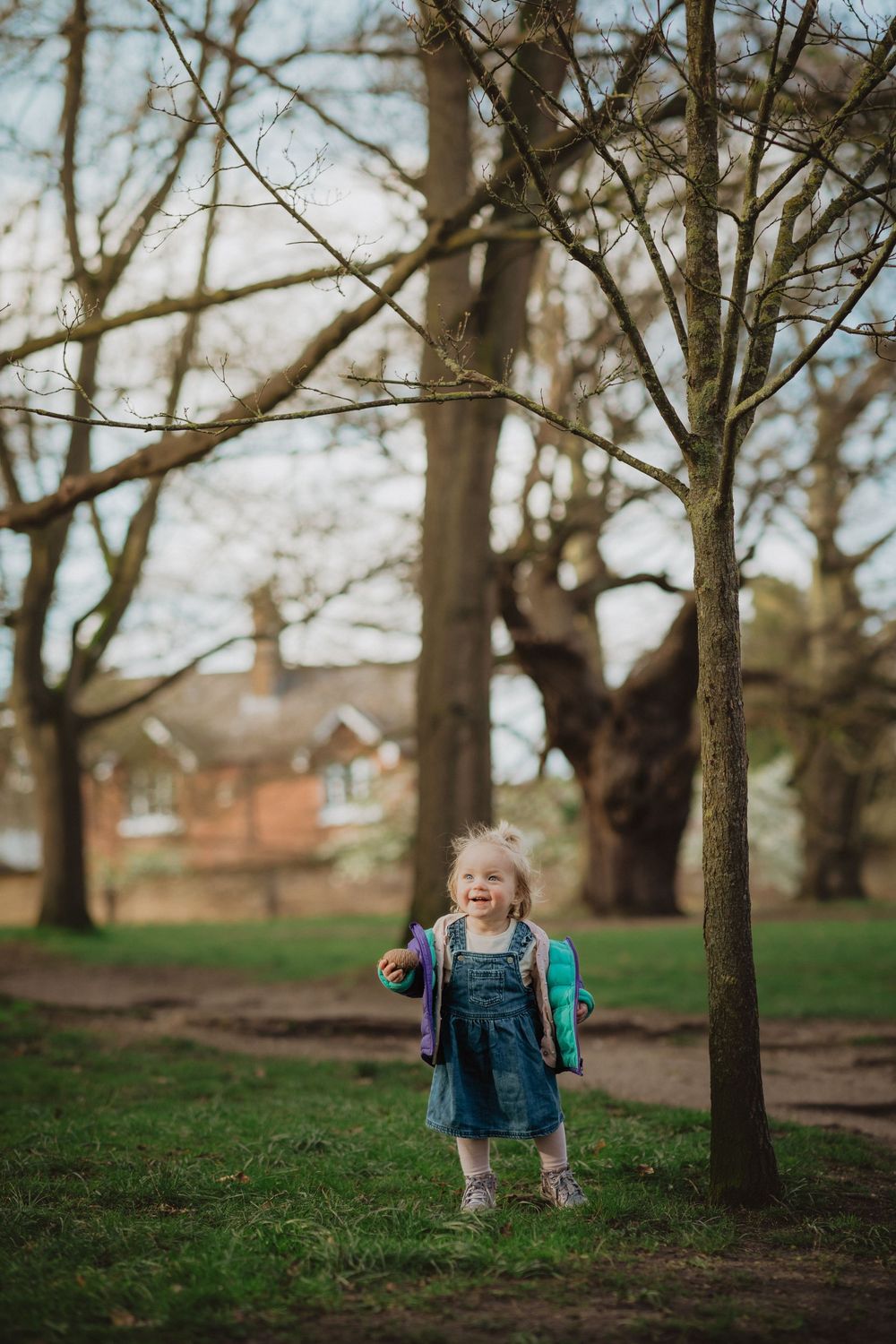 A toddler in overalls and a backpack explores under bare trees near houses.