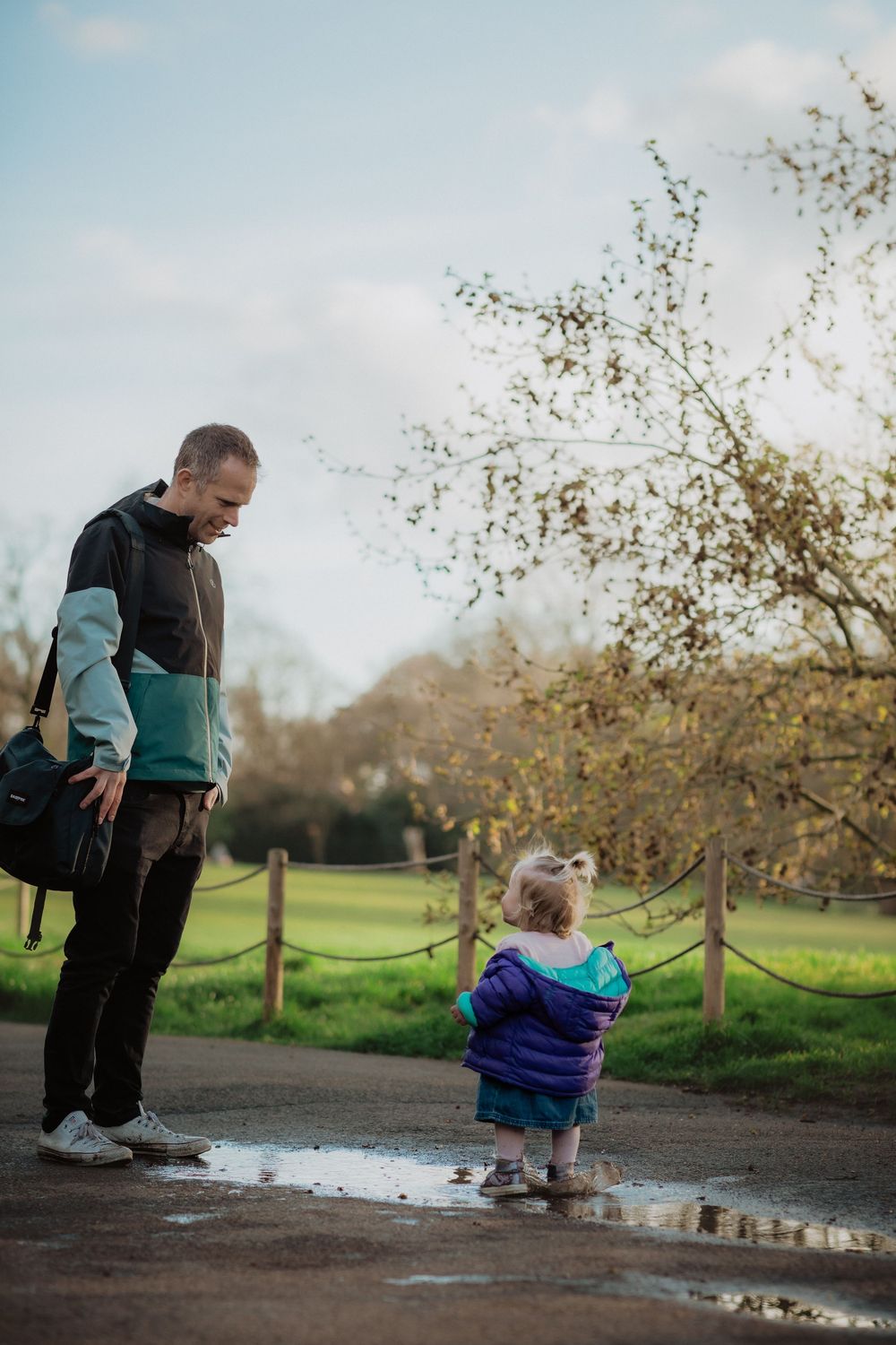 Family exploring a park path together, stopping to look at puddles on a cloudy day.