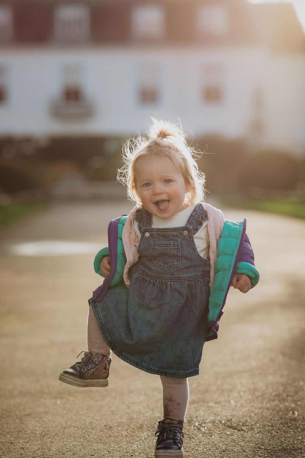 Toddler in denim overalls and backpack skips joyfully on a sunlit path.