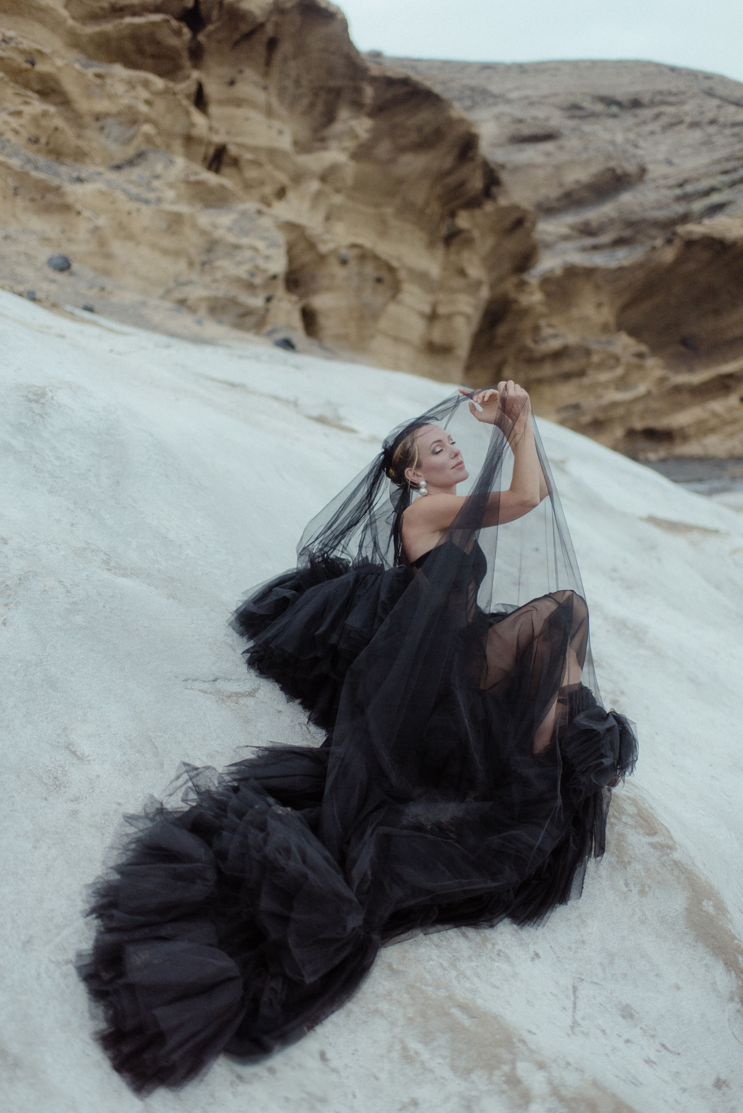 A dramatic figure in a flowing black tulle dress poses gracefully against white sand dunes and rocky desert cliffs in the south of Tenerife.