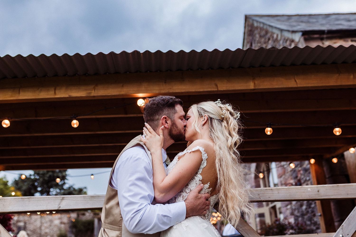 A romantic kiss at a rustic barn venue with string lights glowing overhead at sunset.