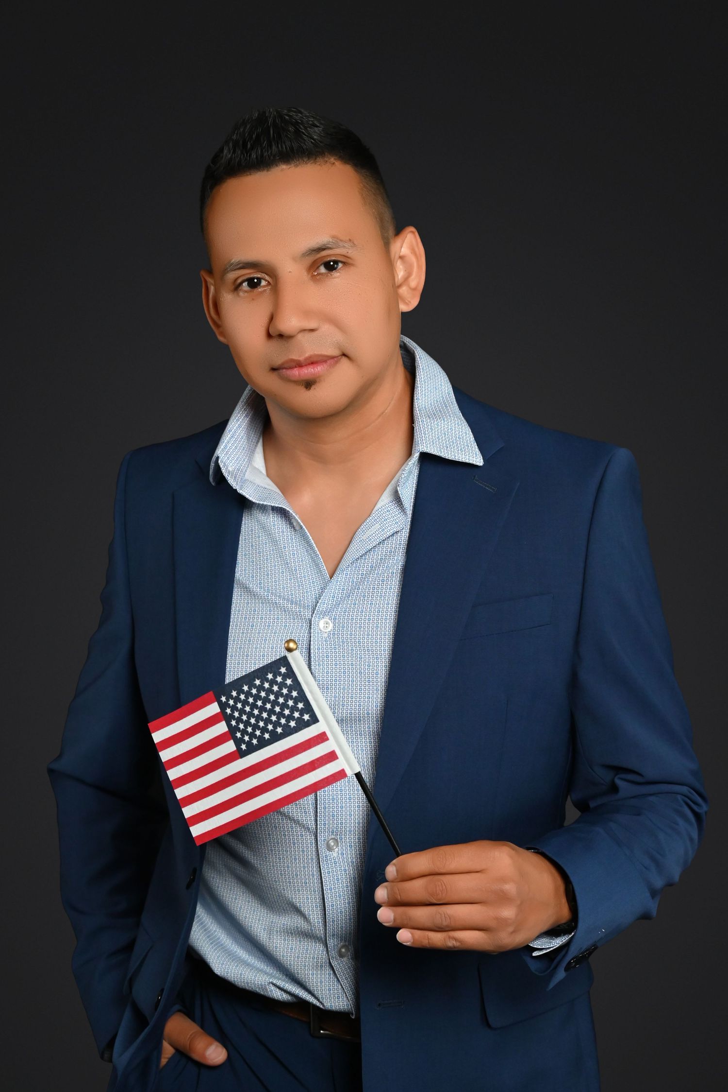 Professional studio portrait of someone in a navy blue blazer and light blue shirt holding a small American flag.