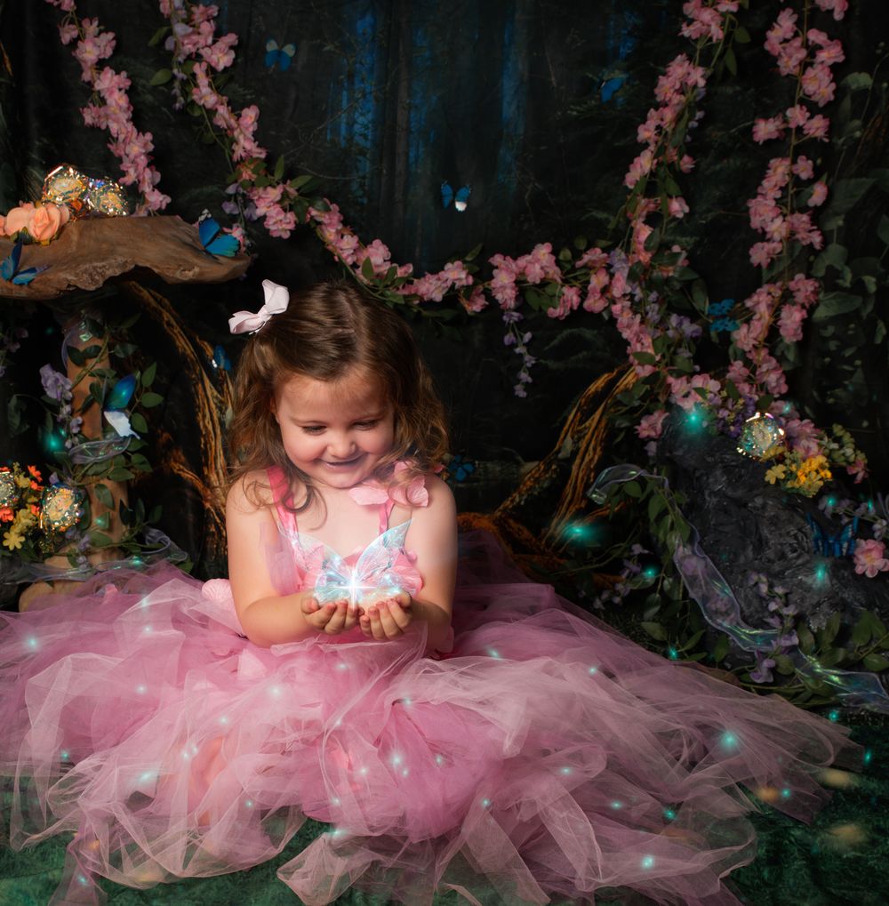 A small child in a pink tulle dress sits with a glowing magical light in an enchanted forest setting with cherry blossoms.
