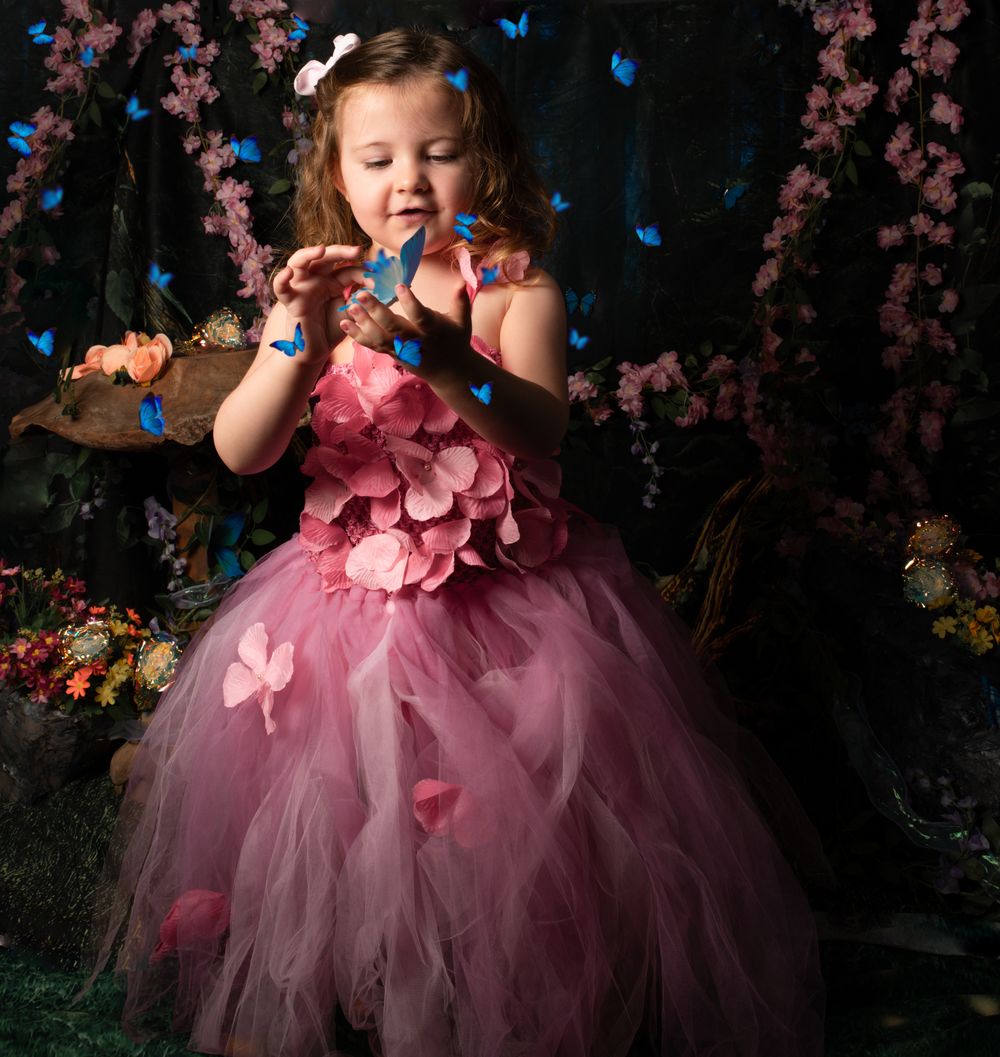 A figure in a pink tulle dress with floral details poses in a magical garden setting with butterflies and cherry blossoms.