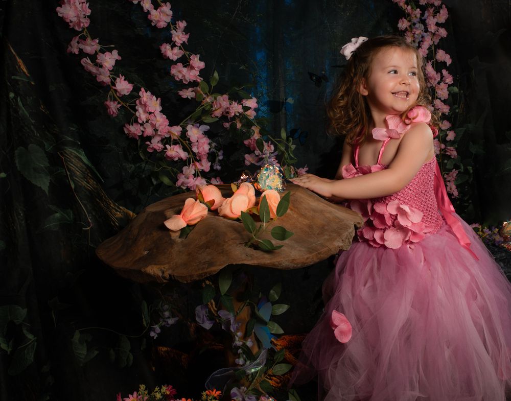 A child in a rosy gown poses beside a wooden table decorated with pink blossoms and whimsical forest elements.